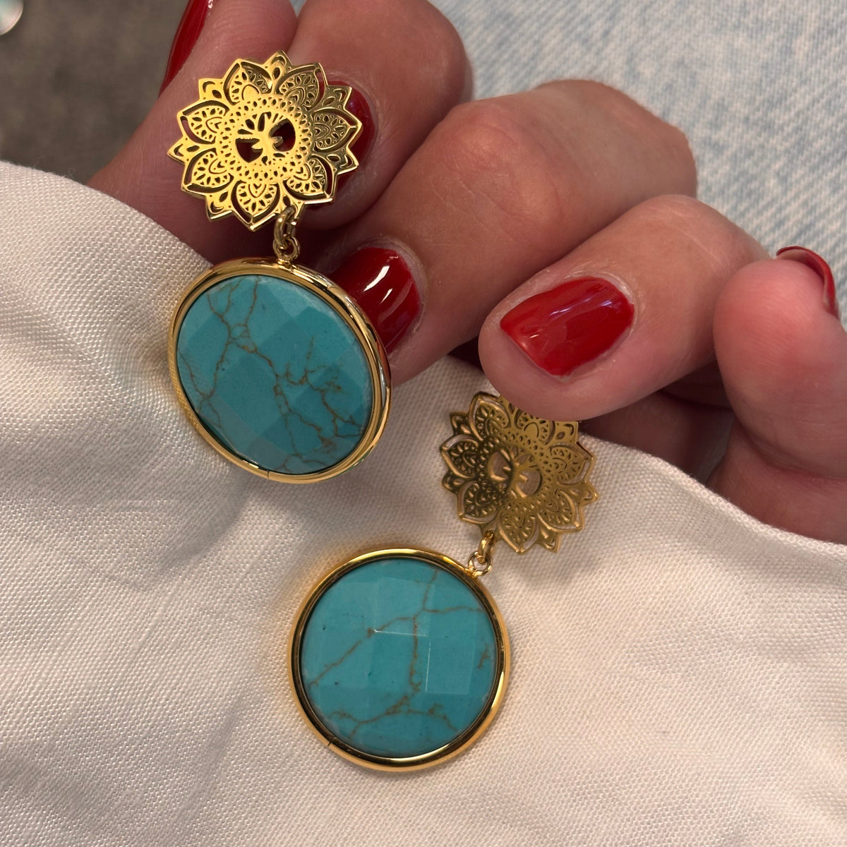 Gold and turquoise earrings held by a hand with red nail polish on a light fabric background.